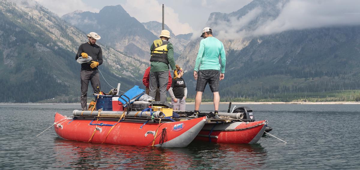 Four researchers prepare for coring on a floating platform on a lake surrounded by high mountains.